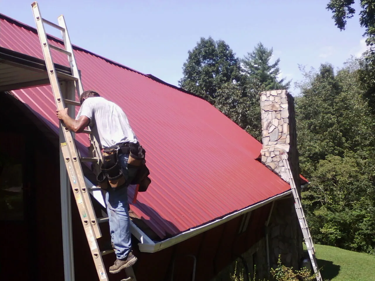 Roofer on a ladder beside a red metal roof and stone chimney — Soffit & Fascia Repair by Roofing Brothers in Port Deposit